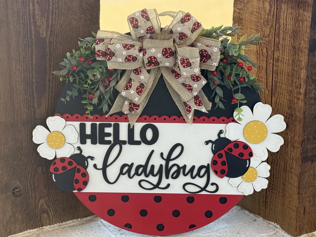 A circular decorative sign with Hello ladybug in black script. It features cartoon ladybugs, white daisies, and red berries. A burlap bow at the top is adorned with red and black ladybugs. The sign has a red and black polka dot base, bordered with green leaves.