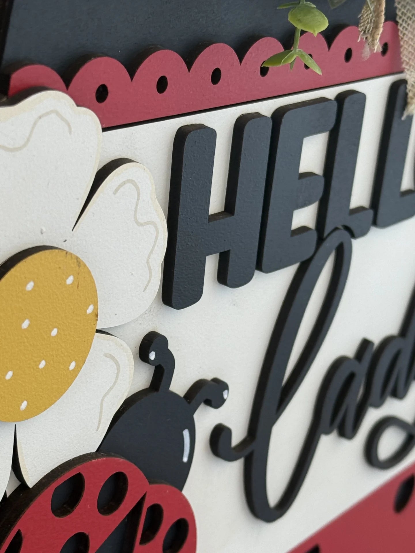 Close-up of a sign with the greeting Hello Lady. The design features a stylized ladybug and a white daisy with a yellow center on a black, white, and red background. The letters are made from thick, cut-out material. A small plant partially obscures the top right corner.