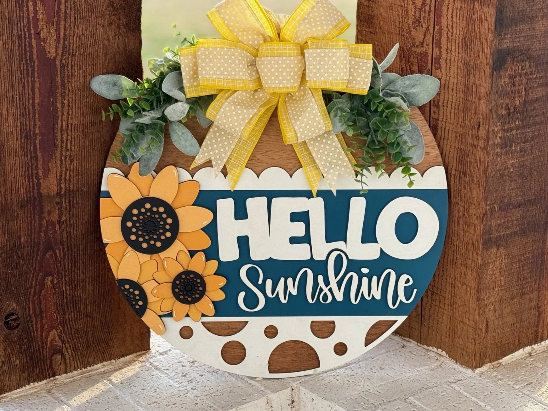 Round decorative wooden sign with Hello Sunshine text. Features yellow sunflowers, a large yellow polka-dotted bow at the top, and green leaves. The background is blue and white, with polka dot accents. It hangs against a wooden backdrop.
