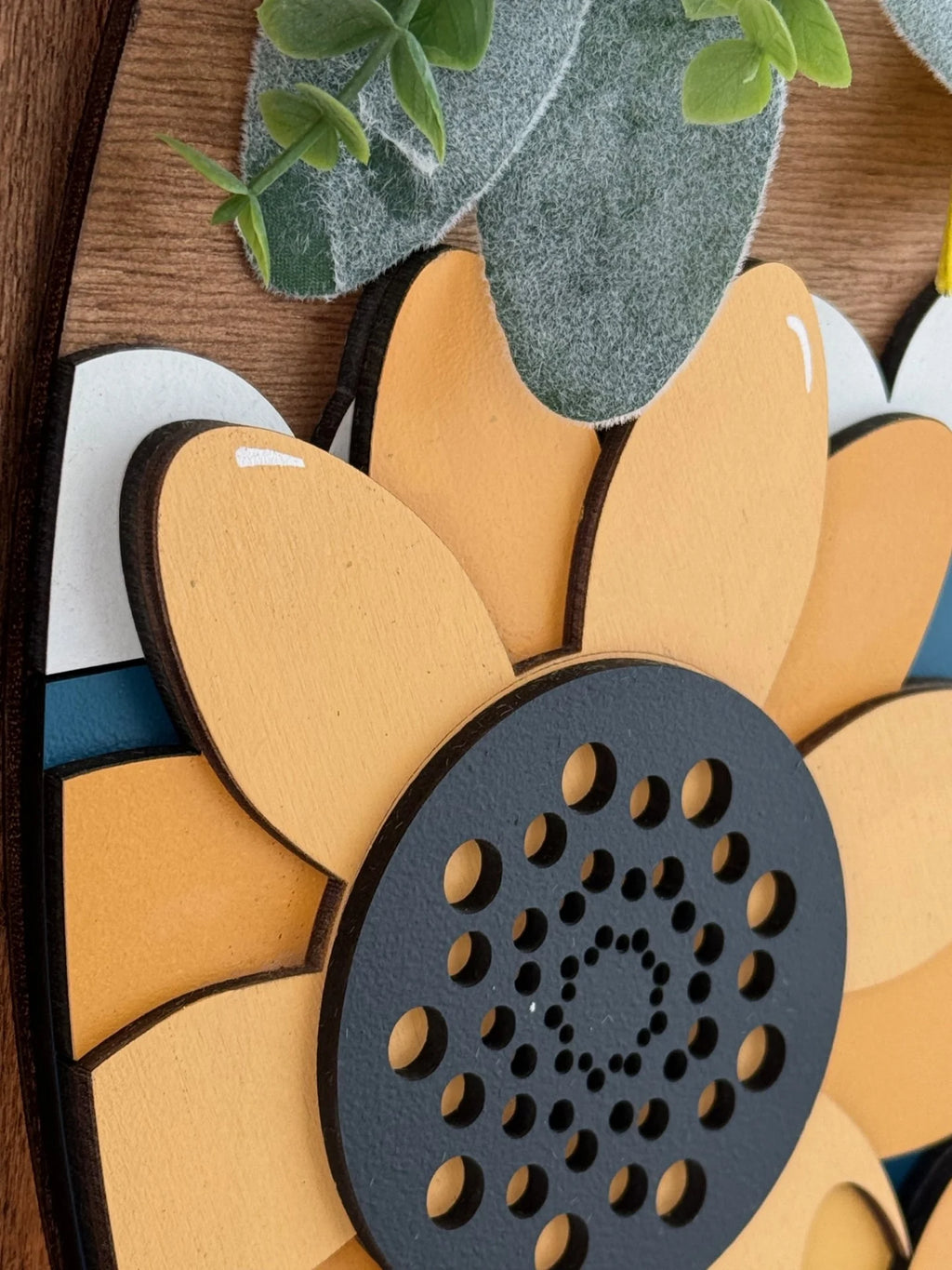 Close-up of a decorative wooden sunflower with orange petals and a dark circular center featuring small holes. Green felt leaves and a textured background accentuate the design, adding a natural touch.