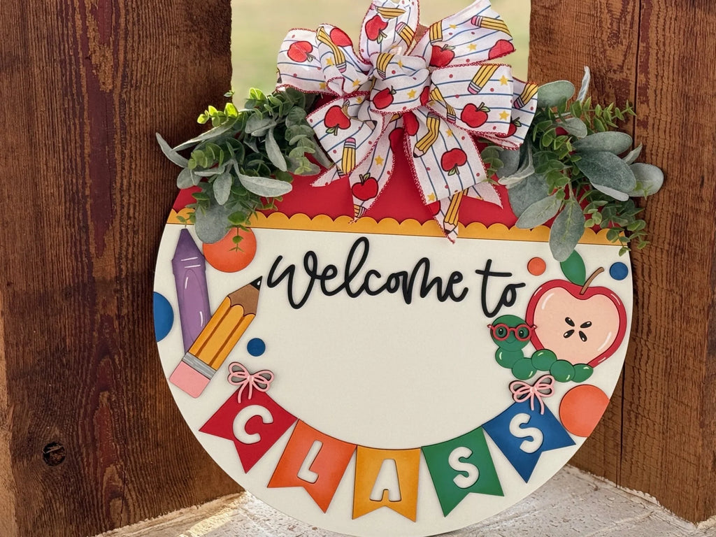 A circular wooden sign with Welcome to Class written in black on a white background. It features colorful school-themed decorations like a pencil, apple, and caterpillar. Topped with a bow adorned with apples and green foliage on a wooden surface. Multicolored banners spell CLASS.