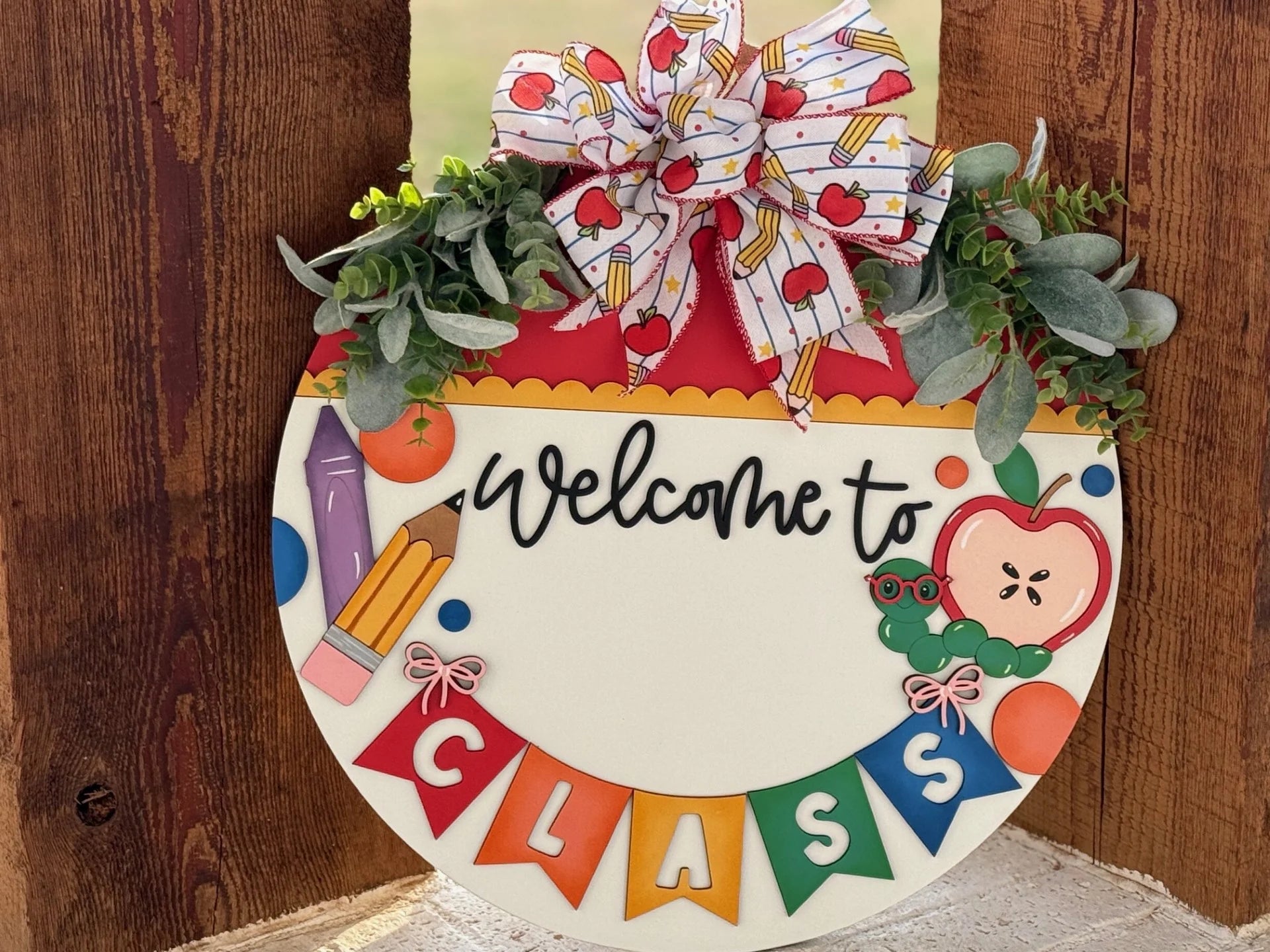A circular wooden sign with Welcome to Class written in black on a white background. It features colorful school-themed decorations like a pencil, apple, and caterpillar. Topped with a bow adorned with apples and green foliage on a wooden surface. Multicolored banners spell CLASS.