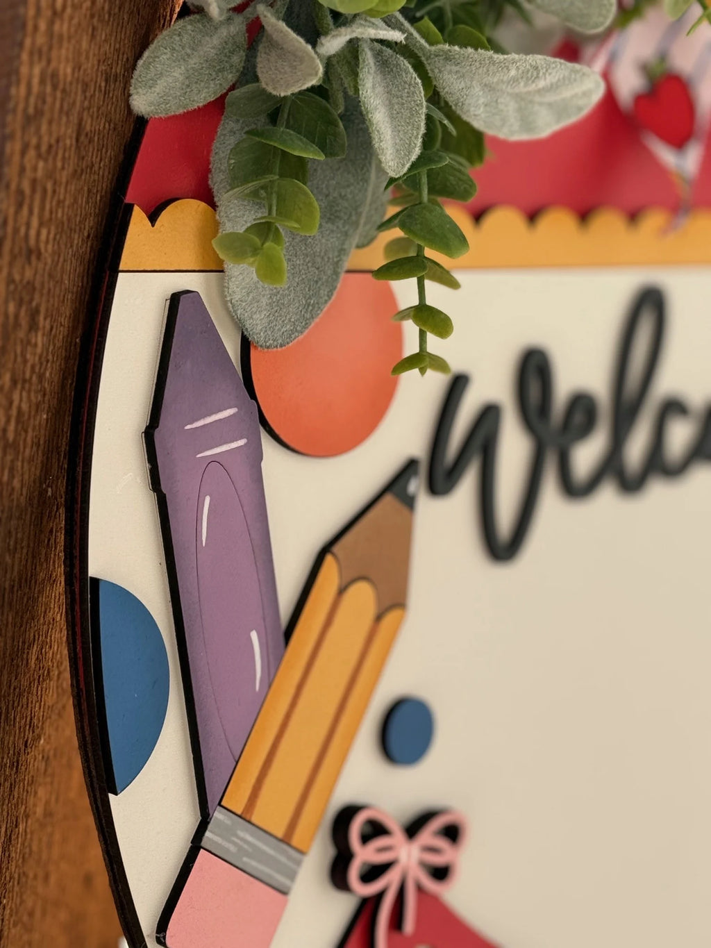 Close-up of a decorative sign featuring a purple pen and a yellow pencil illustration on a multicolored background. The word Welcome is partially visible. Greenery drapes over the top edge, adding a natural touch. The sign is displayed on a wooden surface.