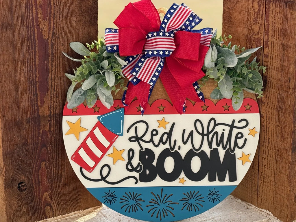 A decorative sign in a half-circle shape features the text Red, White & Boom in black. Its adorned with a red, white, and blue color scheme, stars, fireworks, and a rocket design. A large red bow with stars and stripes is at the top, accented by green leaves, against a wooden background.