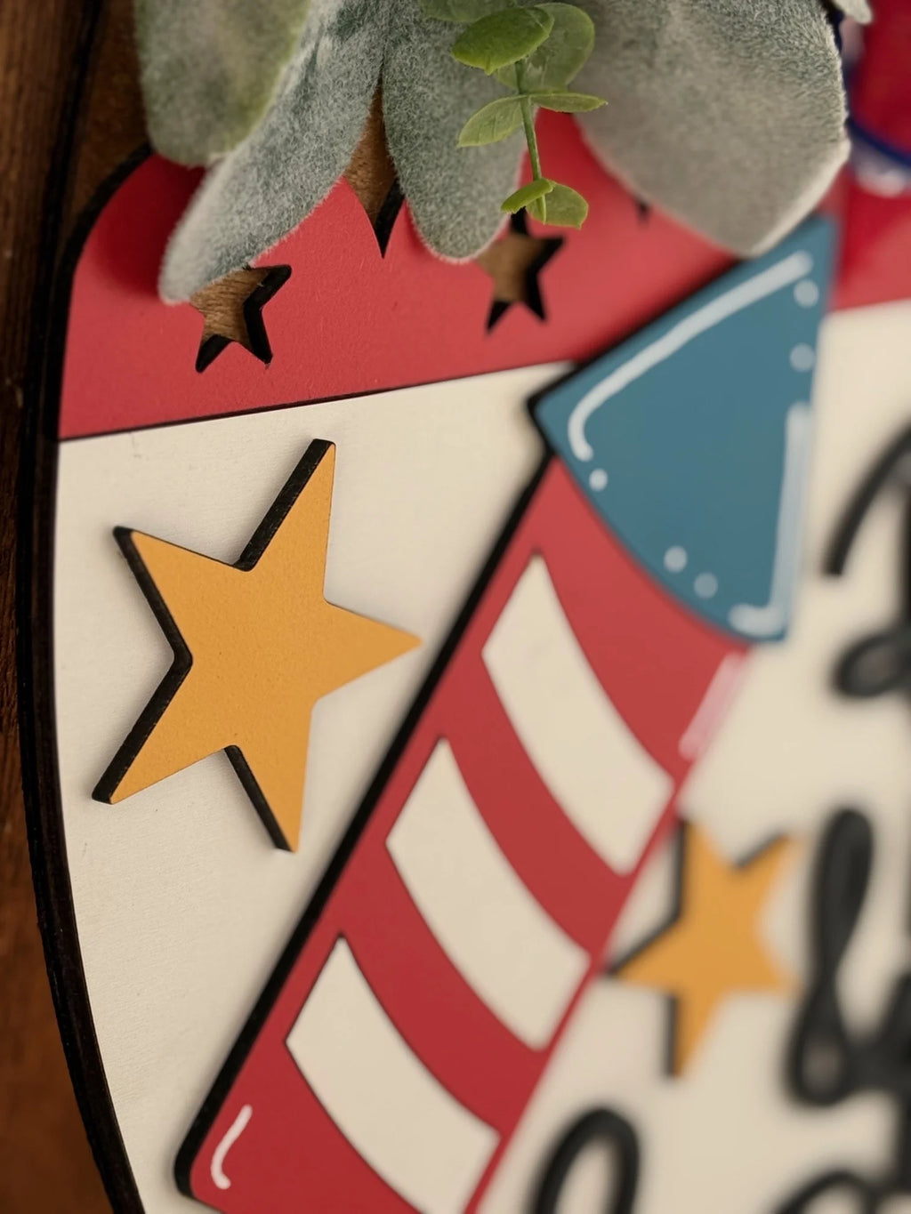 Close-up of a decorative wooden sign featuring a red and white striped firecracker with a blue top, alongside yellow stars. The top of the sign shows greenery, and small black cut-out stars are visible on the red background. The design conveys a festive theme.