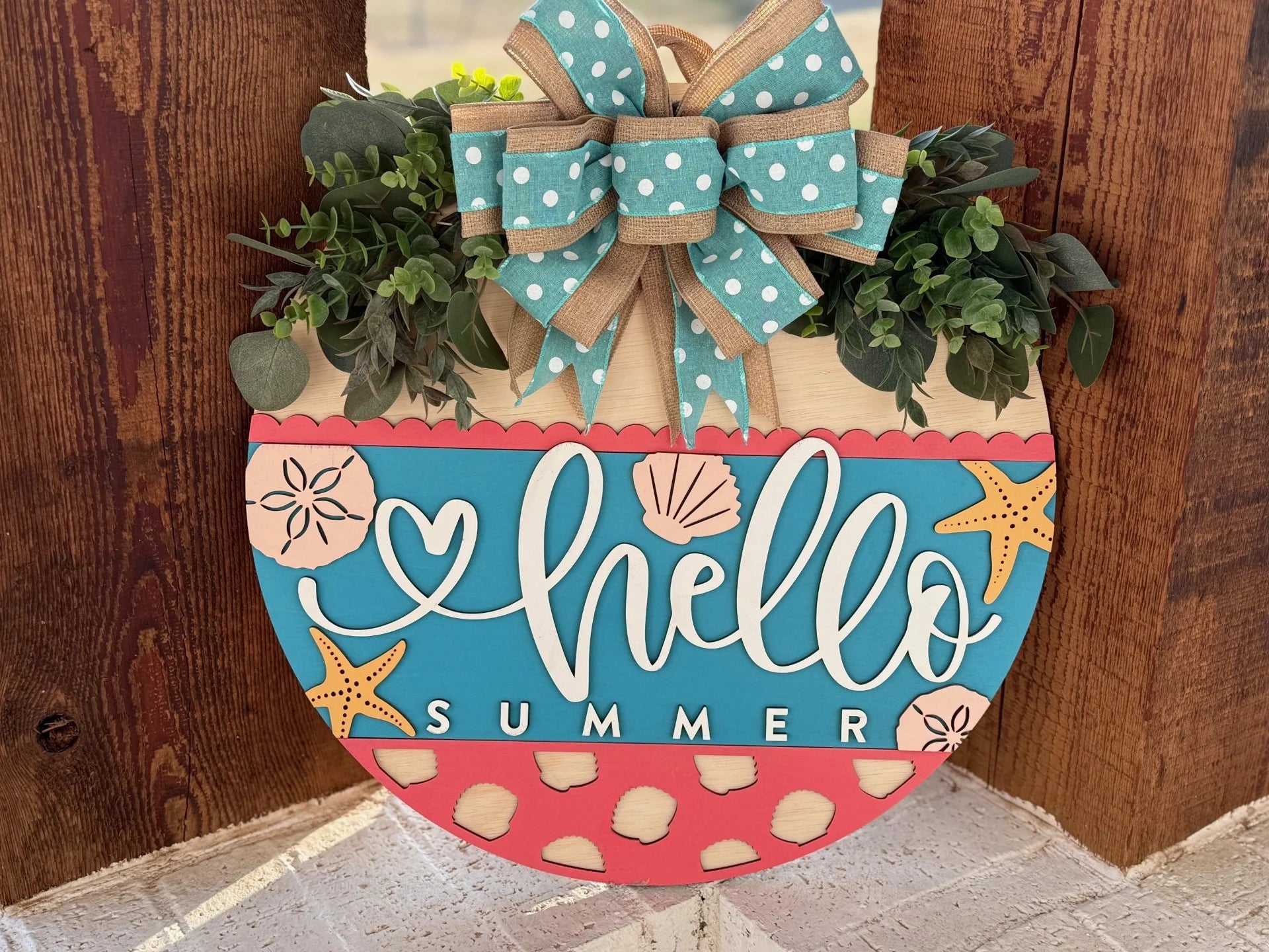 A wooden semi-circular sign with Hello Summer written in white cursive on a blue background. Its adorned with starfish, flowers, and a top border of green leaves. A burlap bow with turquoise and white polka dots sits on top. The bottom is coral with polka dots.