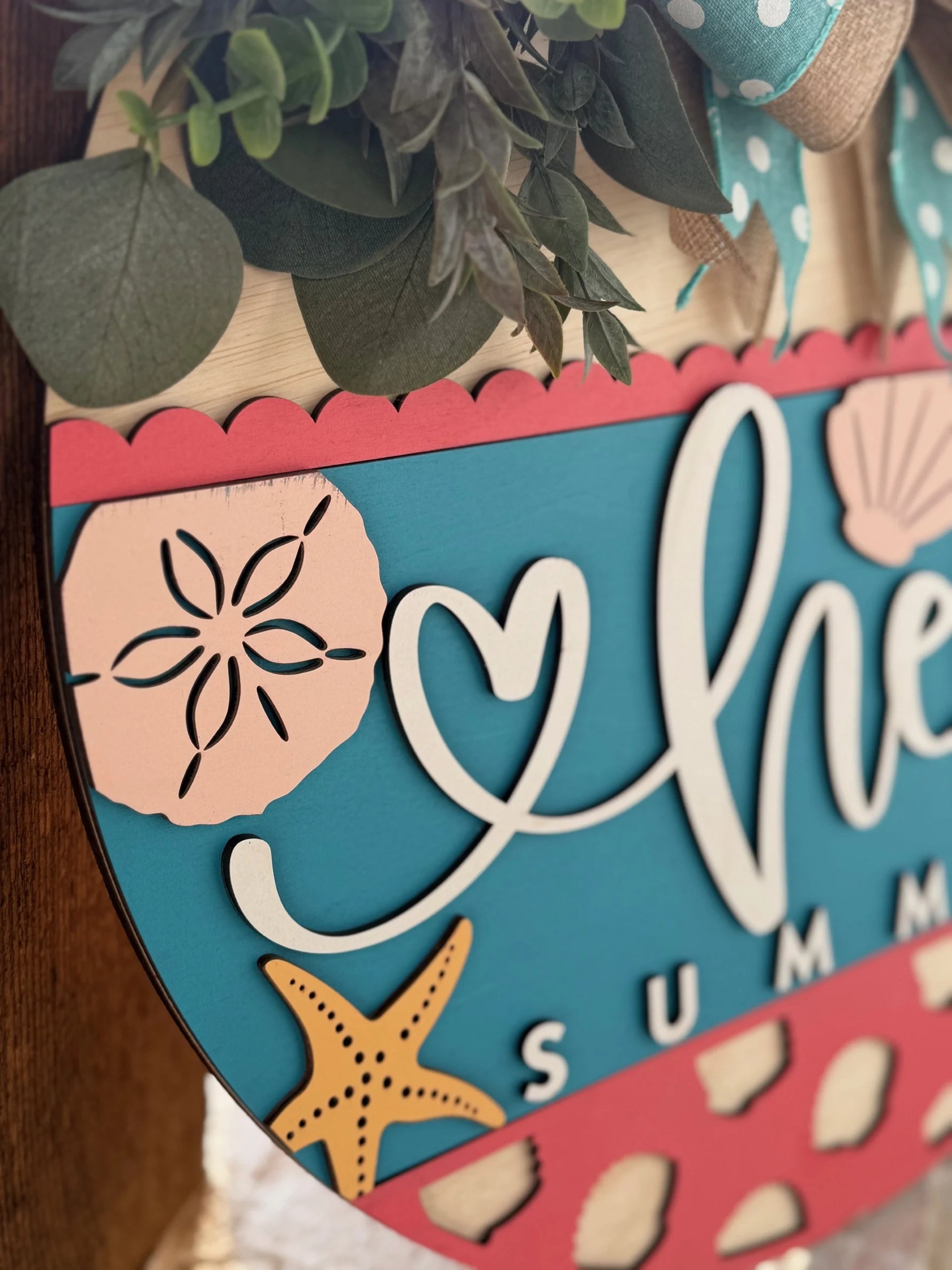 Close-up of a colorful wooden sign with coastal decorations. It features a coral border, teal background, and the word her in white script. Surrounding designs include a pink sand dollar, starfish, and seashell. Greenery and a bow adorn the top.