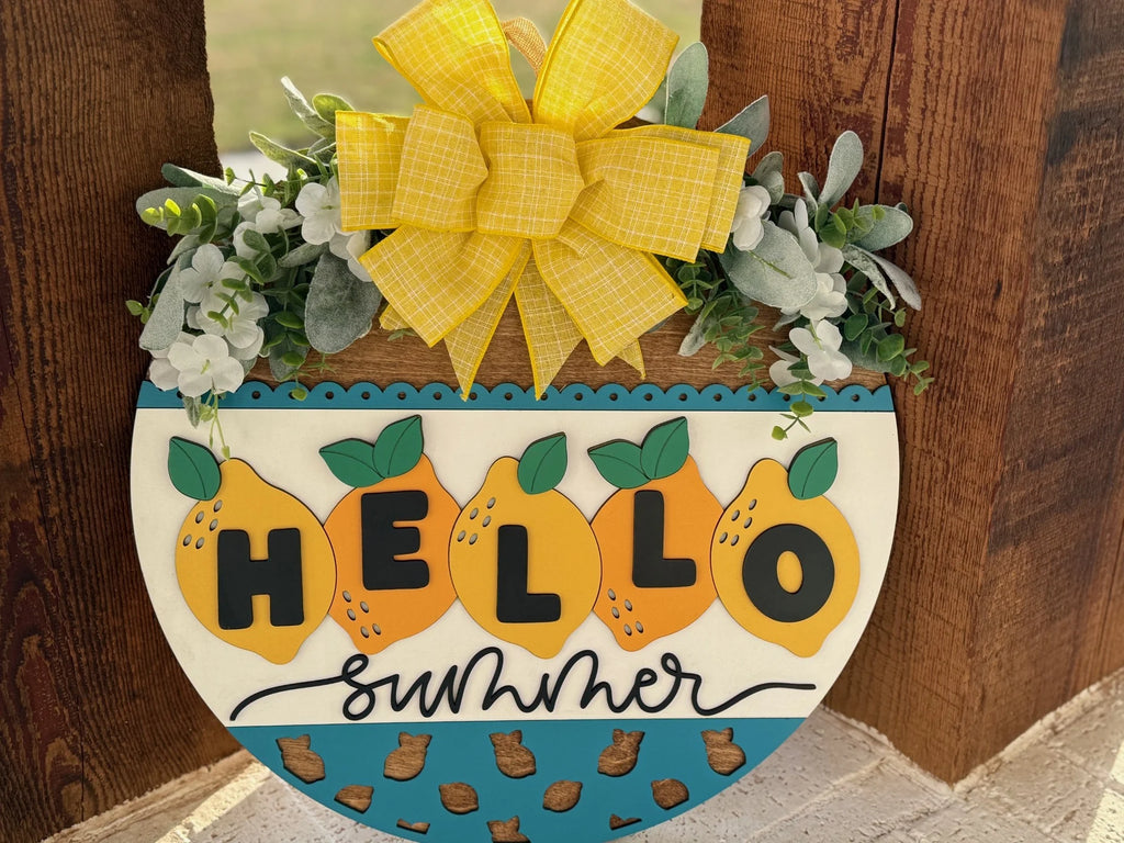 A summer-themed, semi-circular wooden sign features the text Hello Summer with lemon illustrations on a white and blue background. A large yellow bow and green foliage with white flowers adorn the top. The sign is mounted on a rustic wooden surface.