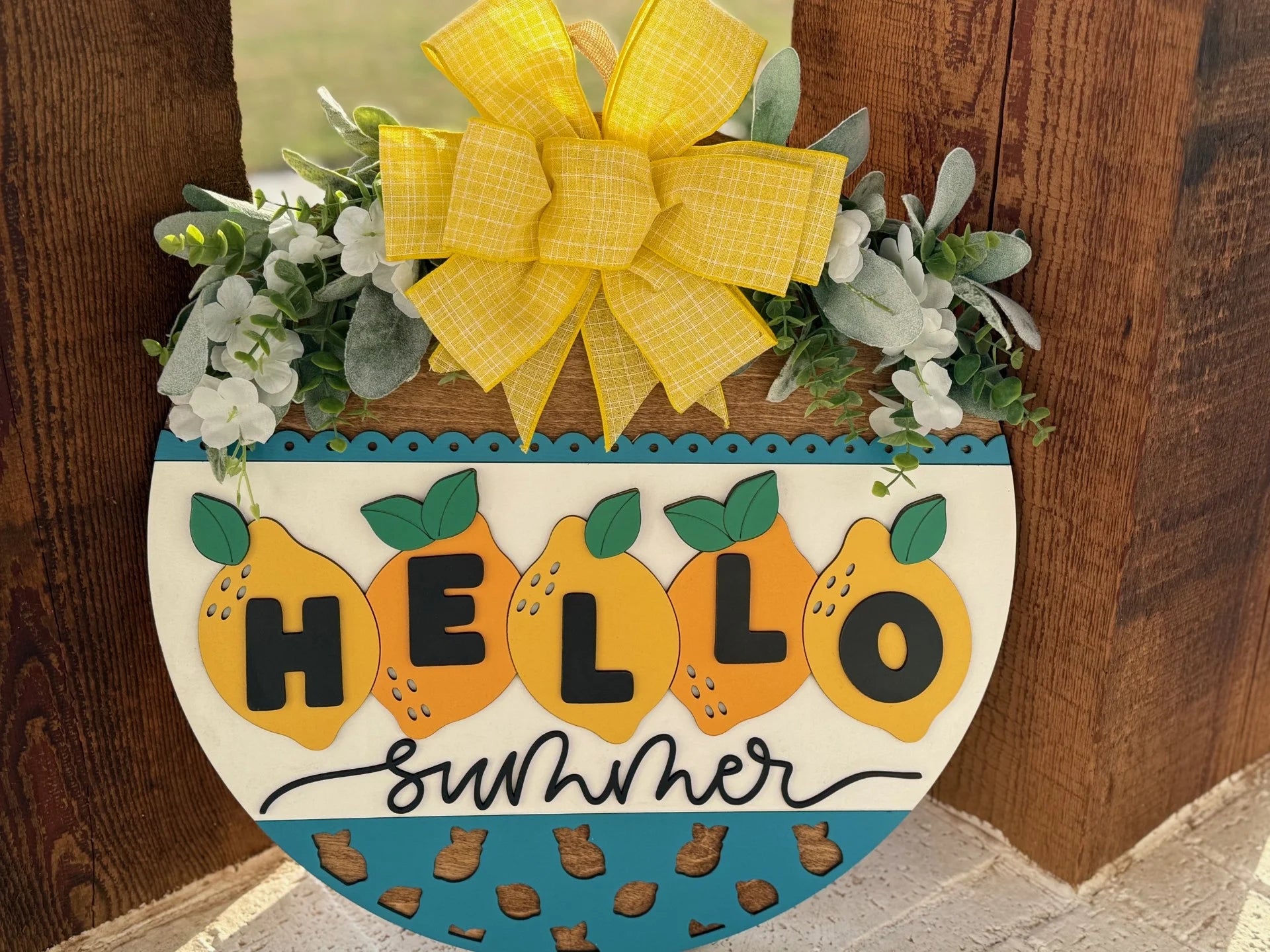 A summer-themed, semi-circular wooden sign features the text Hello Summer with lemon illustrations on a white and blue background. A large yellow bow and green foliage with white flowers adorn the top. The sign is mounted on a rustic wooden surface.