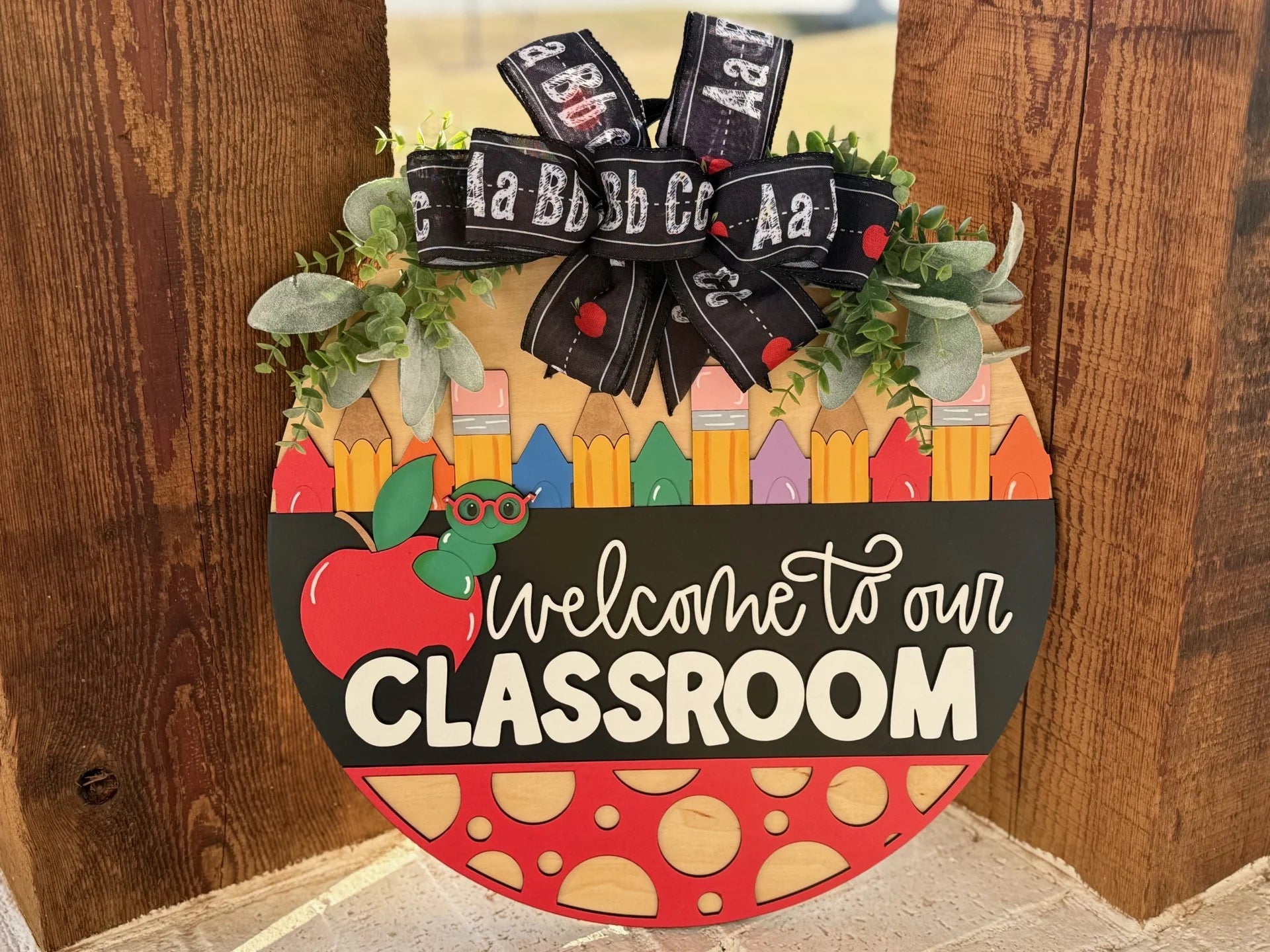 Round wooden sign with colorful houses, foliage, and black ribbon on top. Features red apple and glasses graphics. Text reads, Welcome to our Classroom. Placed against a wooden backdrop.