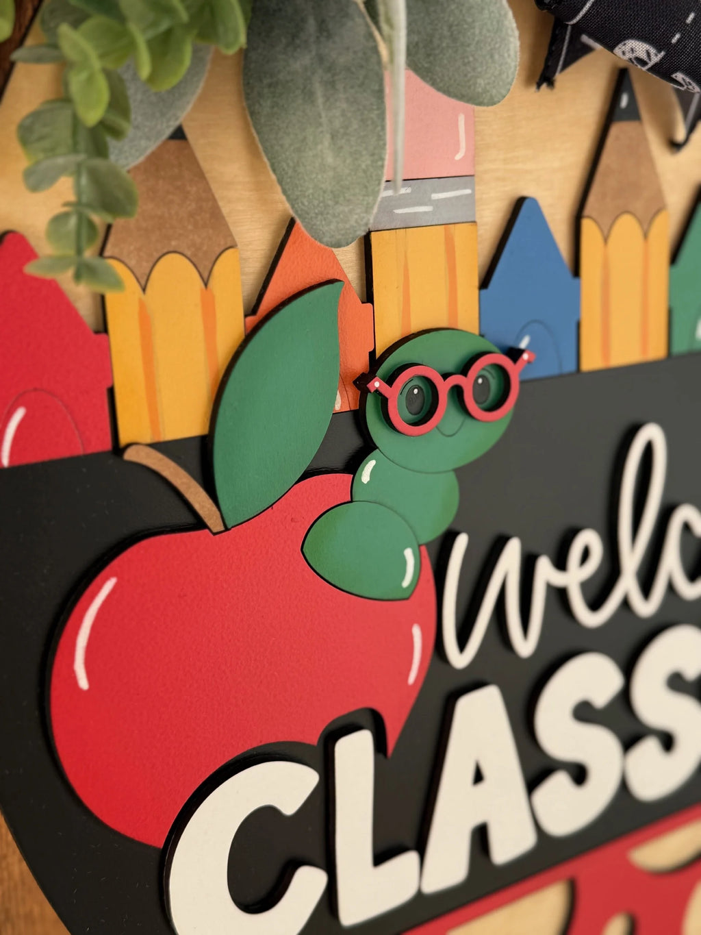 A classroom welcome sign features colorful, pencil-shaped cutouts, a large red apple, and a cute green worm wearing red glasses. Green leaves are at the top. The word Welcome is partially visible against a black background.