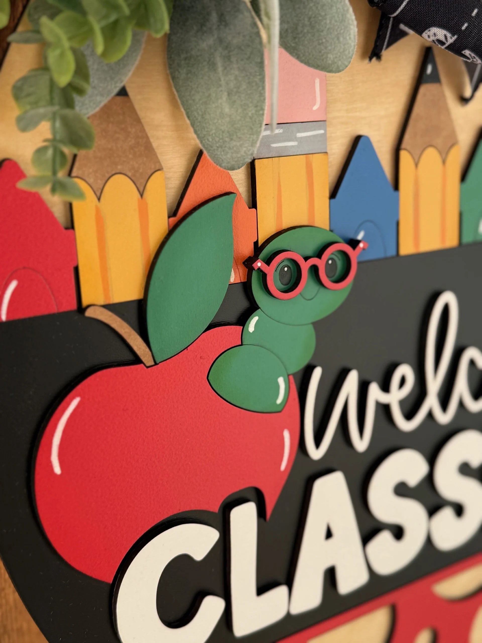 A classroom welcome sign features colorful, pencil-shaped cutouts, a large red apple, and a cute green worm wearing red glasses. Green leaves are at the top. The word Welcome is partially visible against a black background.