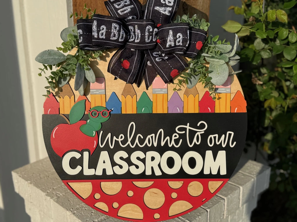 A round, colorful classroom sign on a wall. It features a top half with pencils and a green leaf detail. The center has a chalkboard-style text: Welcome to our Classroom. The bottom has a red apple and cheese pattern. A bow with alphabet letters is attached at the top.