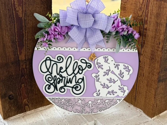 Round wooden sign with Hello Spring in black cursive on a pastel purple background. Features a floral-patterned bunny and lavender flowers with a purple checkered bow on top. Greenery accents and a small butterfly add to the spring theme. Hanging against a wooden backdrop.