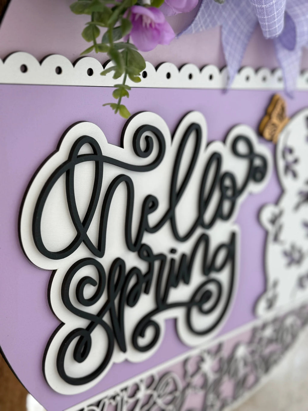 Close-up of a purple sign with the text Hello Spring in swirling black letters, outlined in white. Above, theres decorative greenery and purple flowers. The top edge of the sign is scalloped with a white border. A small brown butterfly embellishment is to the right.