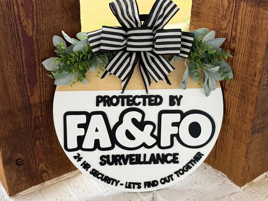 A round wooden sign with a black and white striped bow and green foliage on top. The sign reads: Protected by FA & FO Surveillance. 24 Hr. Security - Lets Find Out Together in bold letters. The sign hangs on a rustic wooden surface.