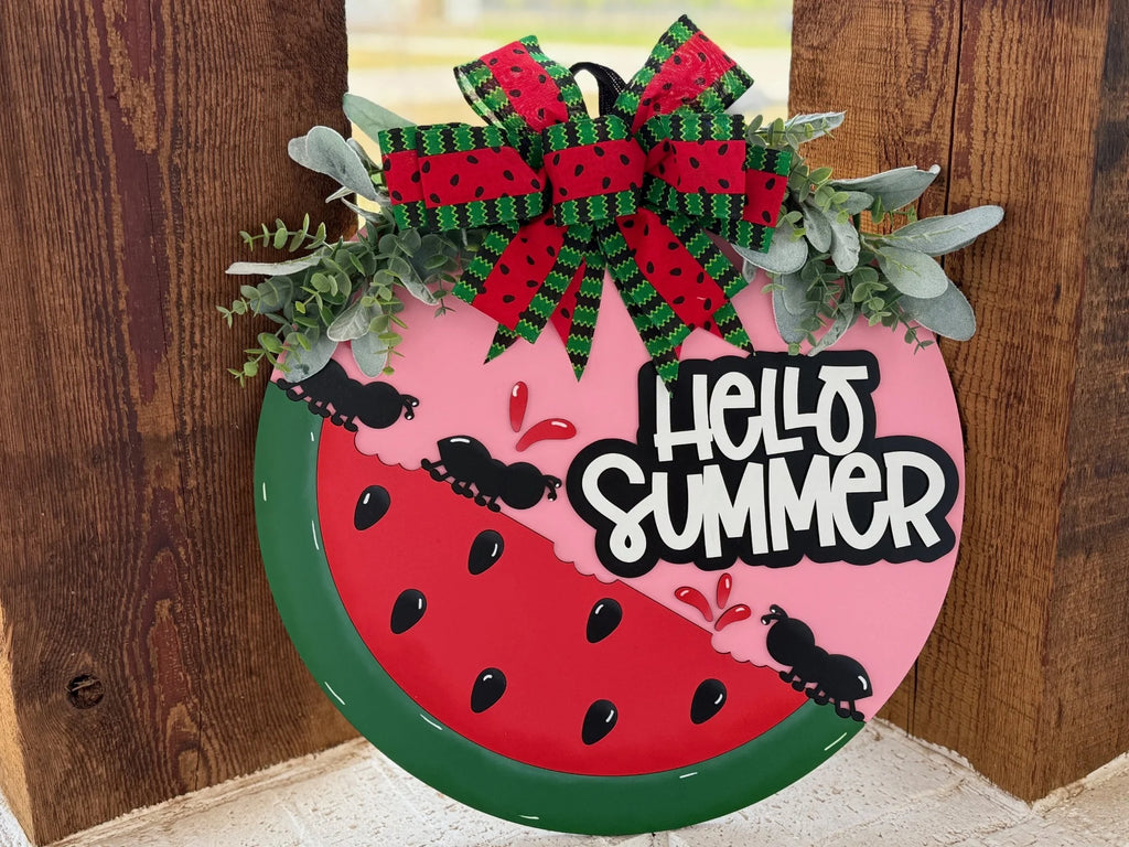 Round decorative sign resembling a watermelon slice with Hello Summer text. It features red and green watermelon-patterned ribbons at the top, green foliage accents, and cartoon ants around tiny watermelon slices. The background shows wooden planks.
