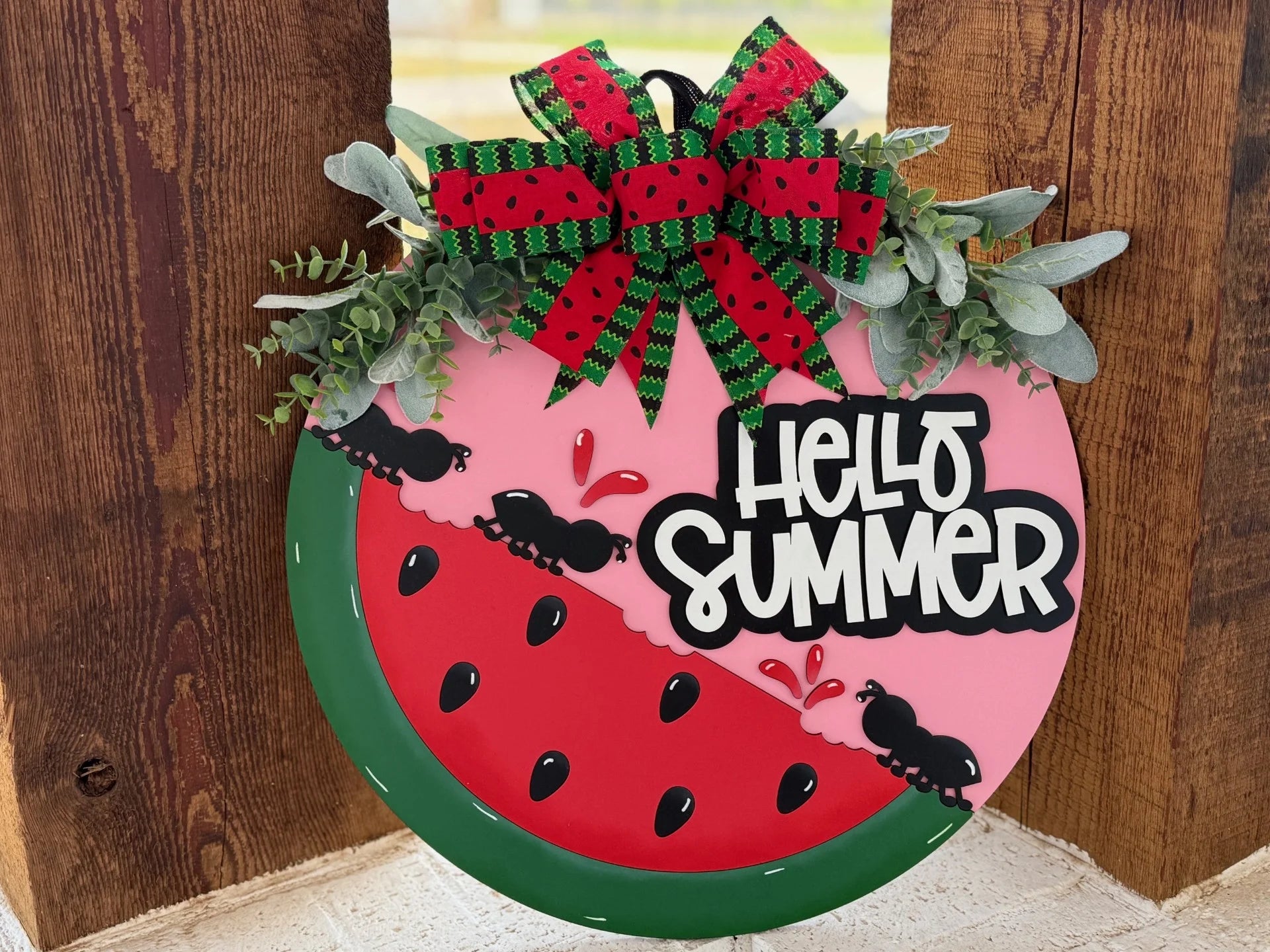 Round decorative sign resembling a watermelon slice with Hello Summer text. It features red and green watermelon-patterned ribbons at the top, green foliage accents, and cartoon ants around tiny watermelon slices. The background shows wooden planks.