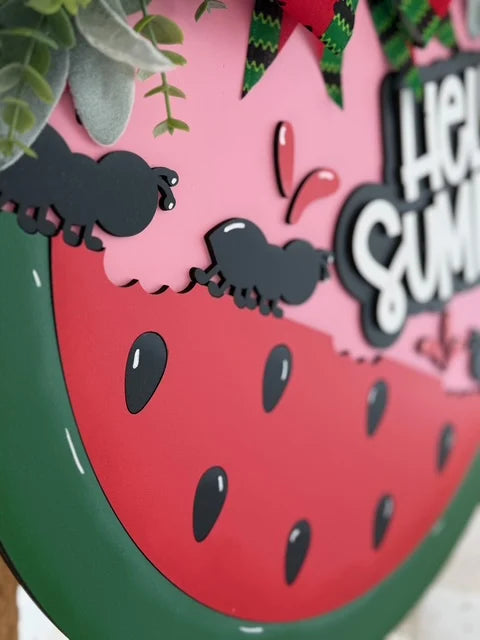 Close-up of a decorative sign featuring a pink and green watermelon slice with black seeds. The top of the sign has cartoon ants carrying food. A red and green bow and leafy garland adorn the corner. The text Hello Summer is partially visible on the right side.
