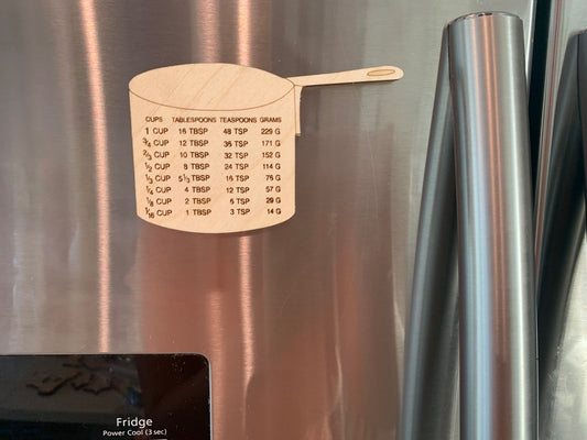 A Kitchen Fridge Magnet Measurements Chart shaped like a pot clings to the stainless steel door, displaying conversions for cups, tablespoons, teaspoons, and grams—ideal for cooking or baking. A handle and display screen are also visible.