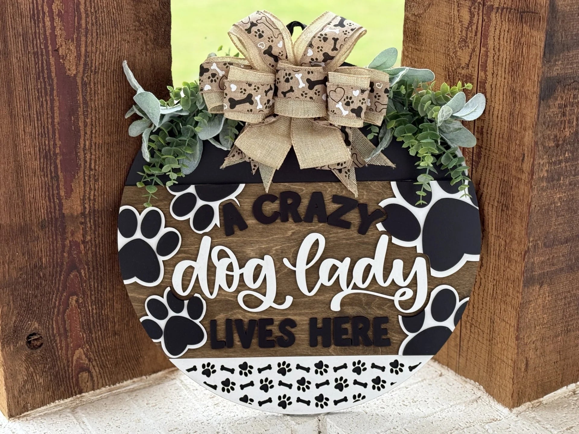 Round wooden sign with the text A CRAZY dog lady LIVES HERE in white. Decorated with black paw prints and a patterned beige bow with dog bones and paws, surrounded by green faux foliage. The sign hangs on a wooden background.