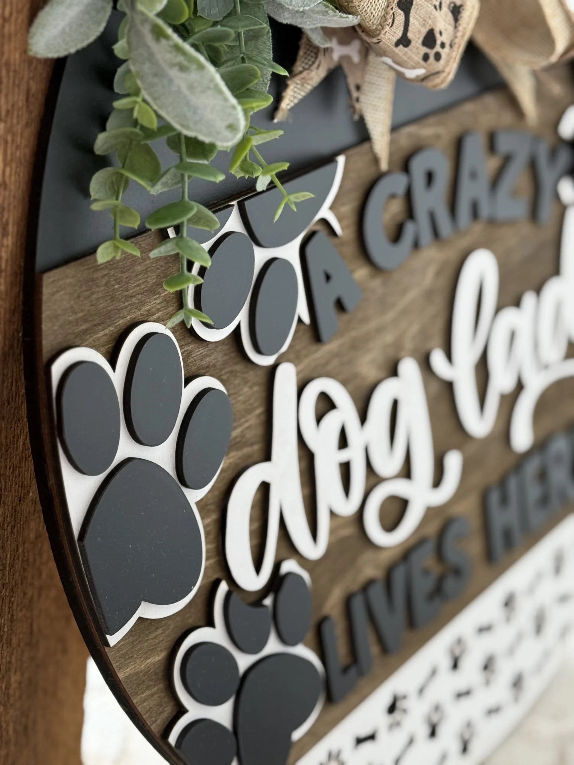 Close-up of a wooden sign with white cursive text reading, A crazy dog lady lives here. The sign features black paw print cutouts and is adorned with green faux eucalyptus leaves and a decorative burlap bow. The background is wooden with a dark edge, giving a rustic appearance.