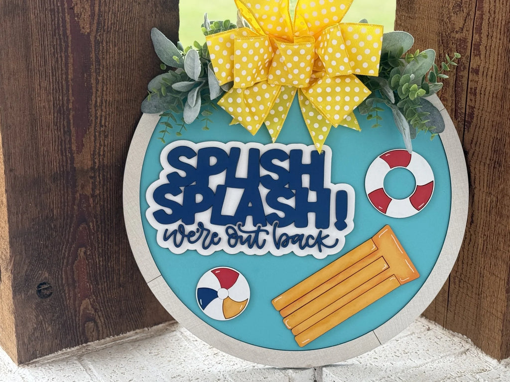 Round door sign with a yellow polka dot bow, greenery, and playful summer items. Features text: SPLISH SPLASH! were out back in blue and white. Includes a beach ball, two lifebuoys, and two pool floats on a blue background, mounted on a wooden door.