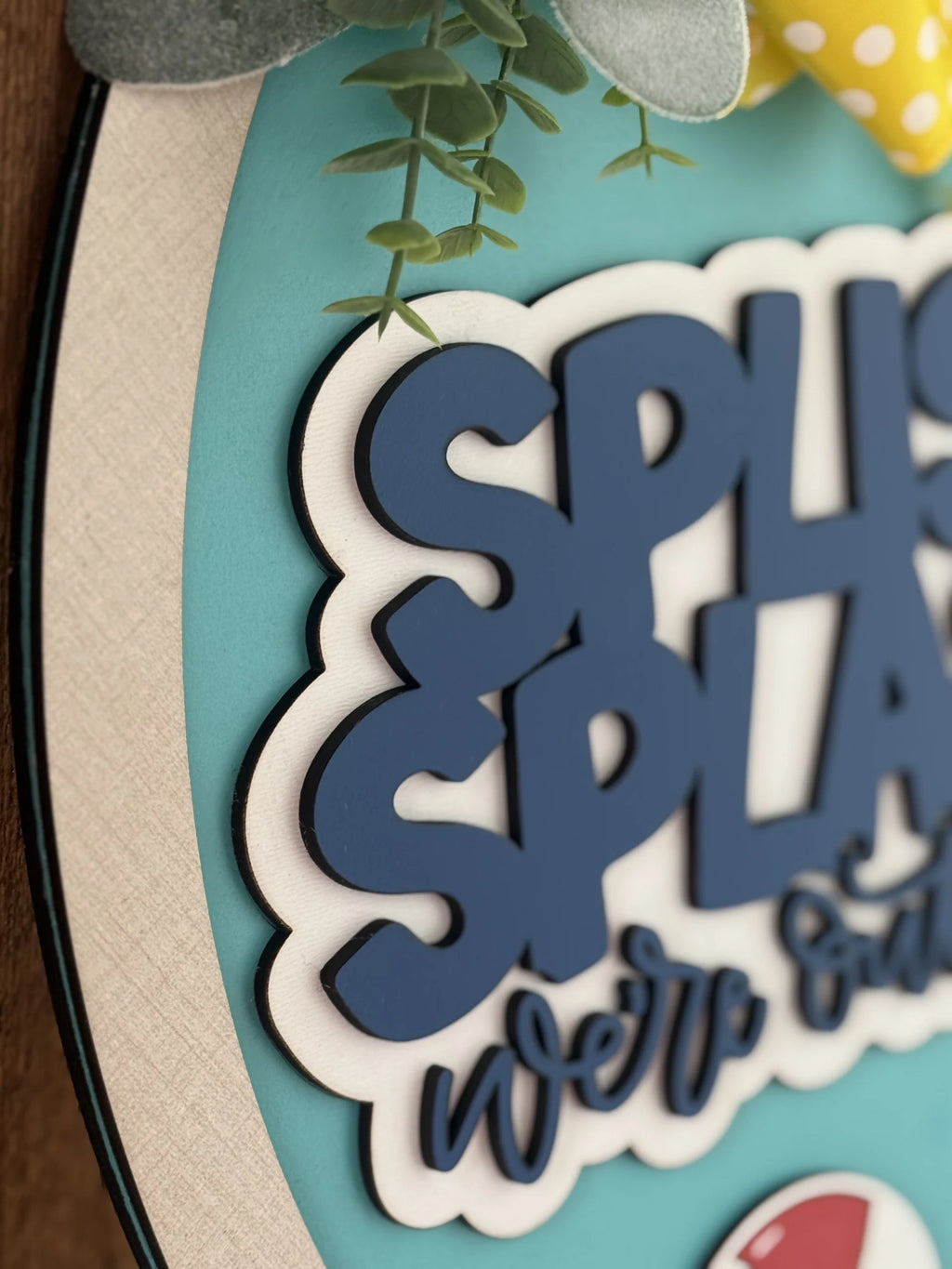 Close-up of a decorative sign with the words SPLISH SPLASH in bold blue letters on a textured teal background. White clouds surround the text. Greenery, a yellow polka dot ribbon, and small decorative elements are visible. The edge is framed by a white border.
