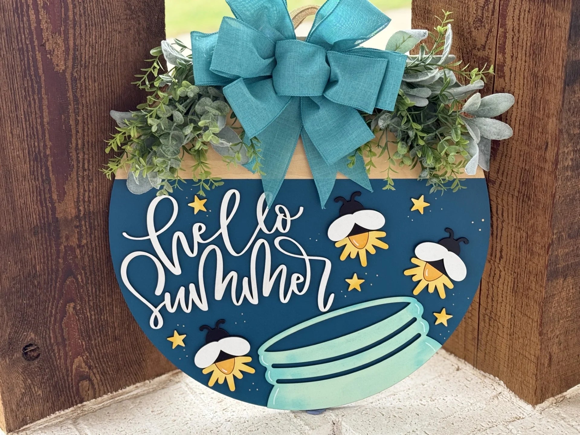 Round wooden sign with a teal background, featuring the text Hello Summer in cursive. Decorated with a large blue bow, greenery, cartoon fireflies, and stars above a mason jar at the bottom. The sign is set against a wooden backdrop.