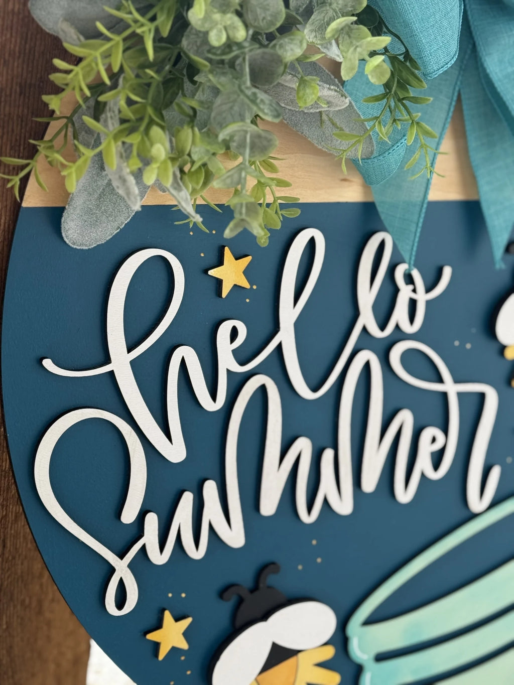 A decorative sign with the phrase Hello Summer in white, curly script on a blue background. The sign features small yellow stars, a bee, and a light blue bow. Greenery is arranged at the top for accent, adding a fresh, summery touch.