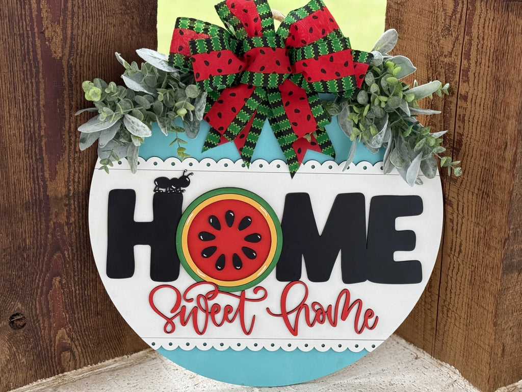 A decorative sign with the word HOME in bold black letters. The O is a red watermelon slice. Below, red cursive text reads sweet home. Atop the sign is a red and green plaid bow with greenery and two ladybugs, all set against a wooden background.