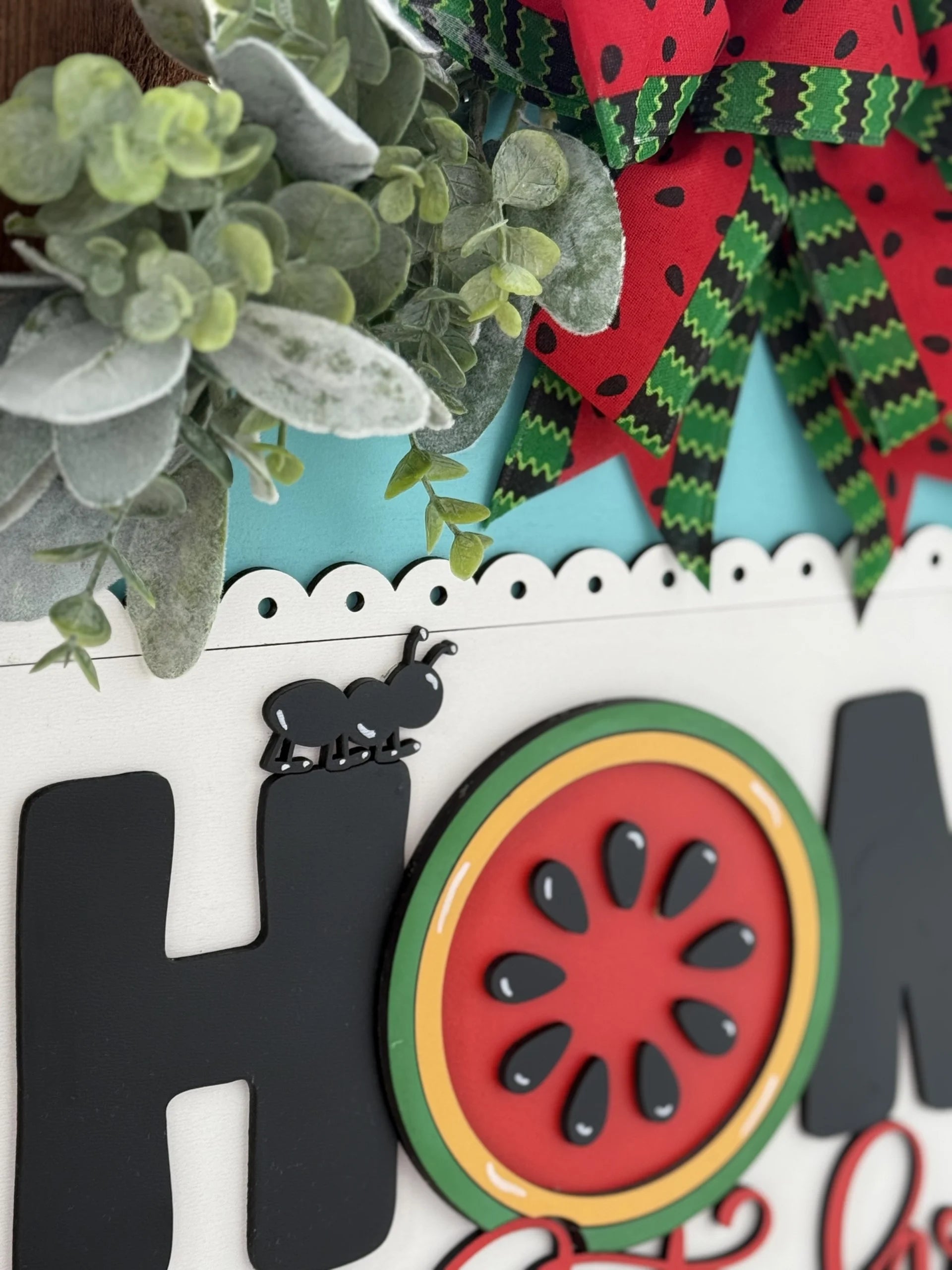 A decorative sign with a floral arrangement on top left and a large, colorful bow in red and green. The word HOME is displayed with a black ant on the H and a watermelon design as the O. The background features a light blue, scalloped edge.