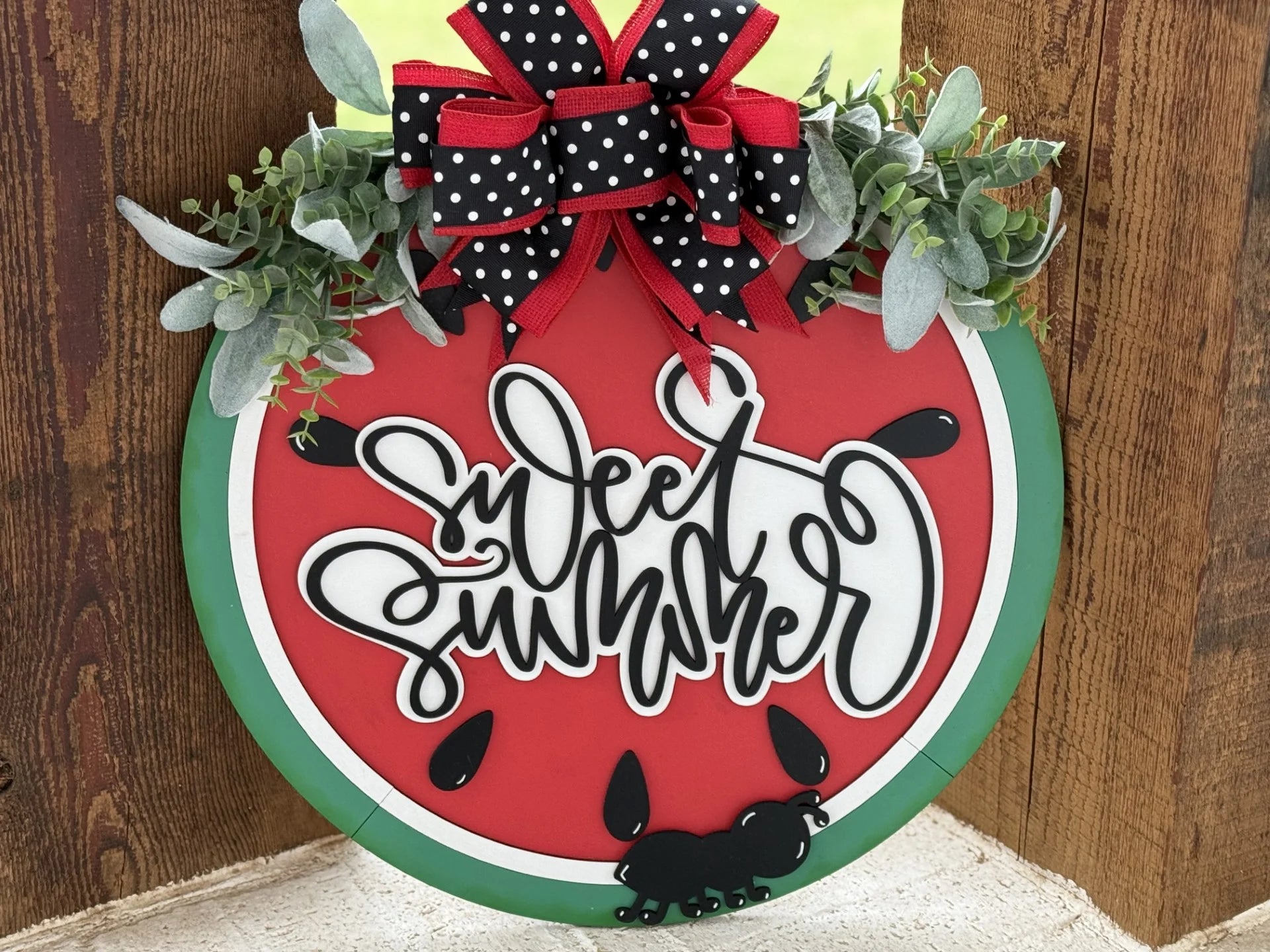 A round decorative sign resembles a watermelon slice with the words Sweet Summer in stylish black script. Topped with a red and black polka dot bow and green leaves, it features a black ant design. Mounted against a wooden background.