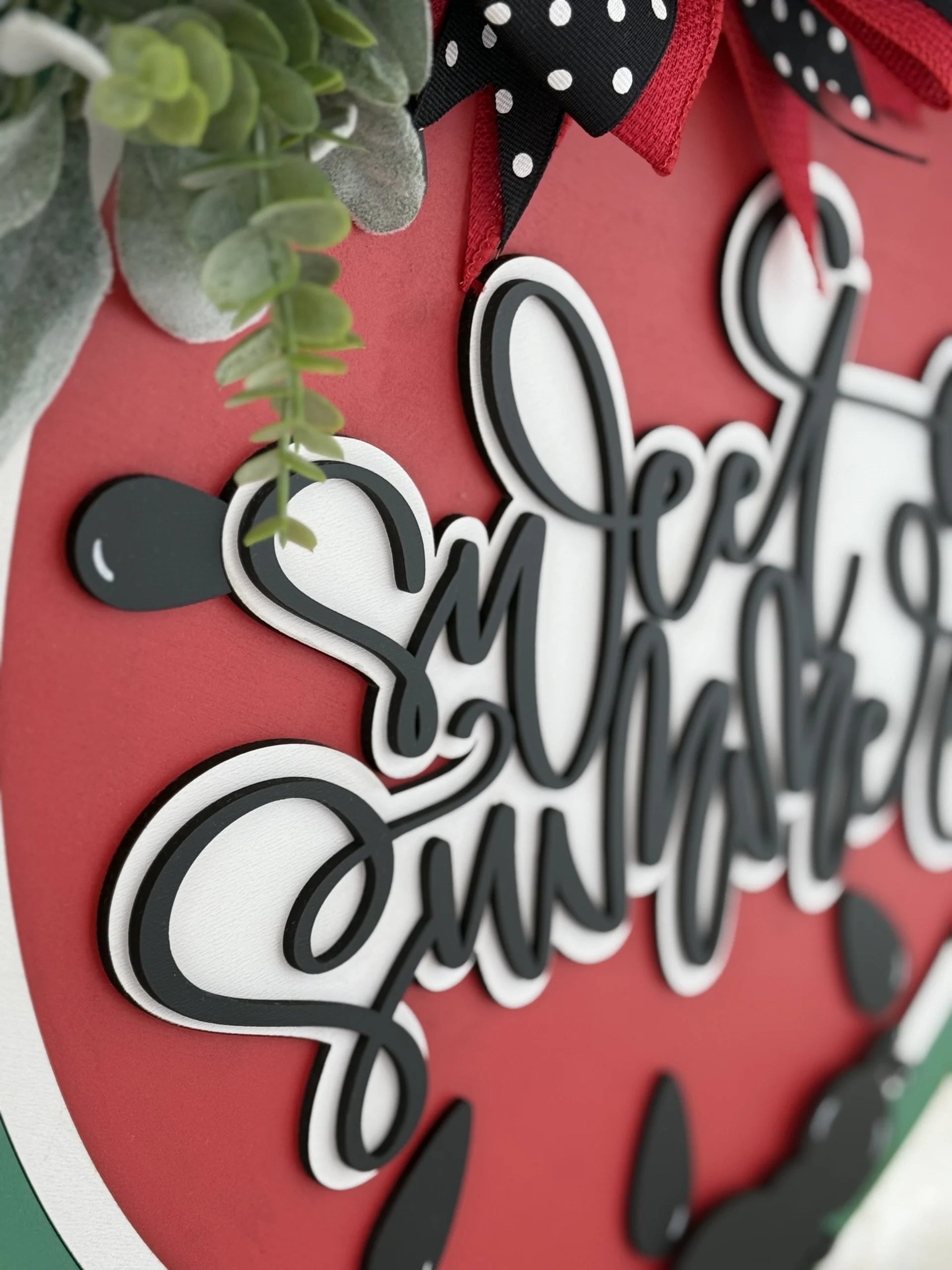 Close-up of a decorative sign featuring a red, watermelon-themed background with black seeds. The script text reads Sweet Summertime in black, layered over the melon design. Greenery and a red and black polka-dotted ribbon adorn the top, adding a festive touch.