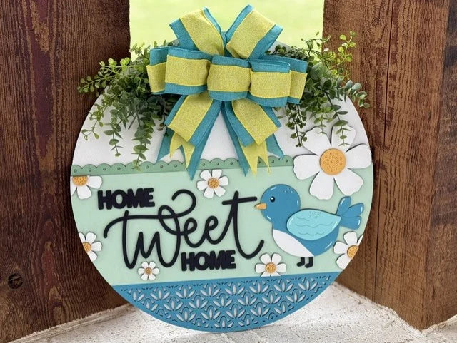 Round decorative sign with “Home Tweet Home” written on it. Features a blue bird, white daisies, and green foliage. A large bow with blue and yellow stripes sits atop. The sign has a light blue patterned lower border and is on a wooden background.