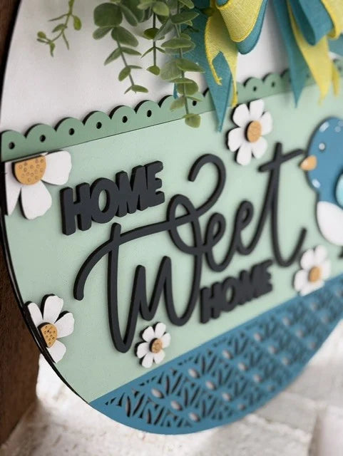 Close-up of a decorative sign featuring the phrase Home Tweet Home. It has a teal background with white daisies, a blue bird, and a yellow bow. The word Tweet is prominently styled in cursive, while Home and Home are in block letters. Greenery decorates the top.