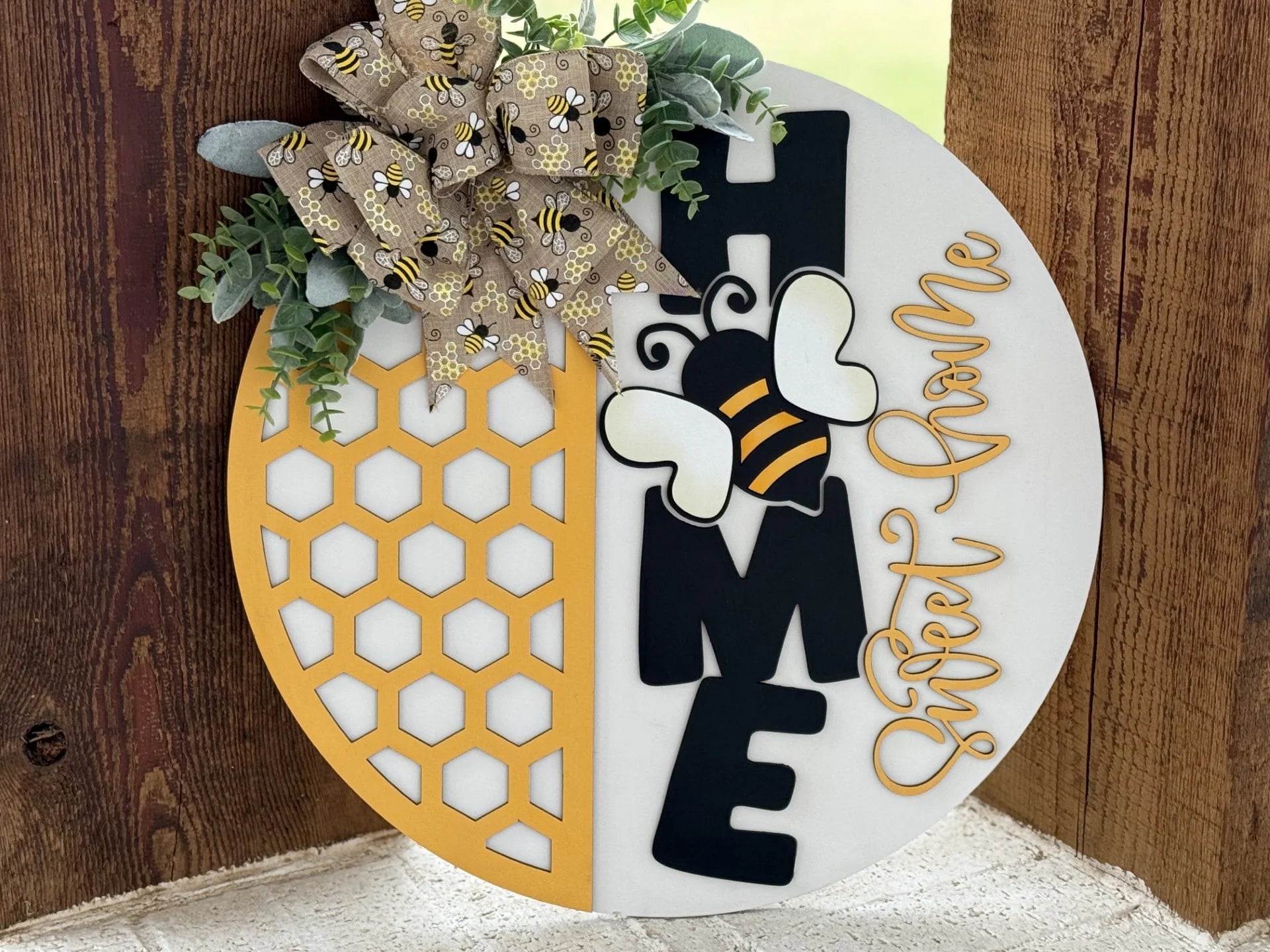 A round, decorative wooden sign displays Home Sweet Home with Home in large black letters and Sweet in yellow script. A cute bee and honeycomb pattern adorn the left half. A bow with bee prints and greenery decorates the top, leaning against a wooden surface.