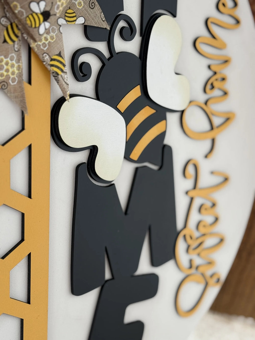 Close-up of a decorative sign featuring a stylized bee with black and yellow stripes. The bee is adorned with white wings and curly antennae. To the left, a honeycomb pattern and a bow with a bee design. Cursive text on the right includes the words “messy” and “welcome.”.