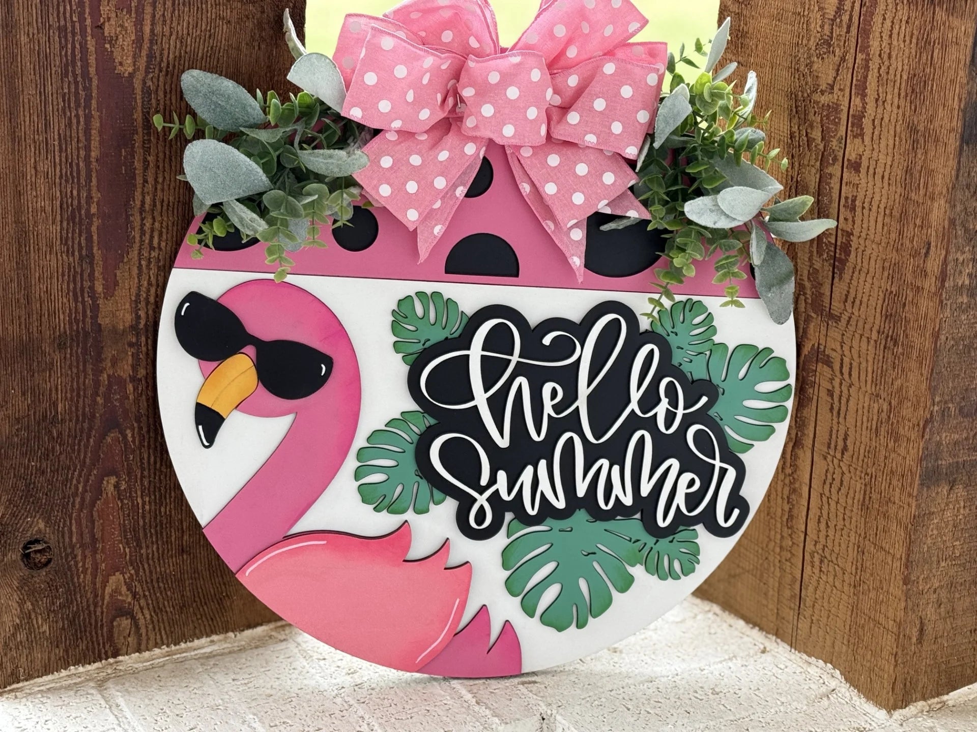 Round wooden sign with a pink flamingo wearing sunglasses and a large pink polka dot bow on top. Surrounded by green leaves, the sign reads hello summer in white cursive. The background is a wooden surface, and the overall theme is bright and tropical.