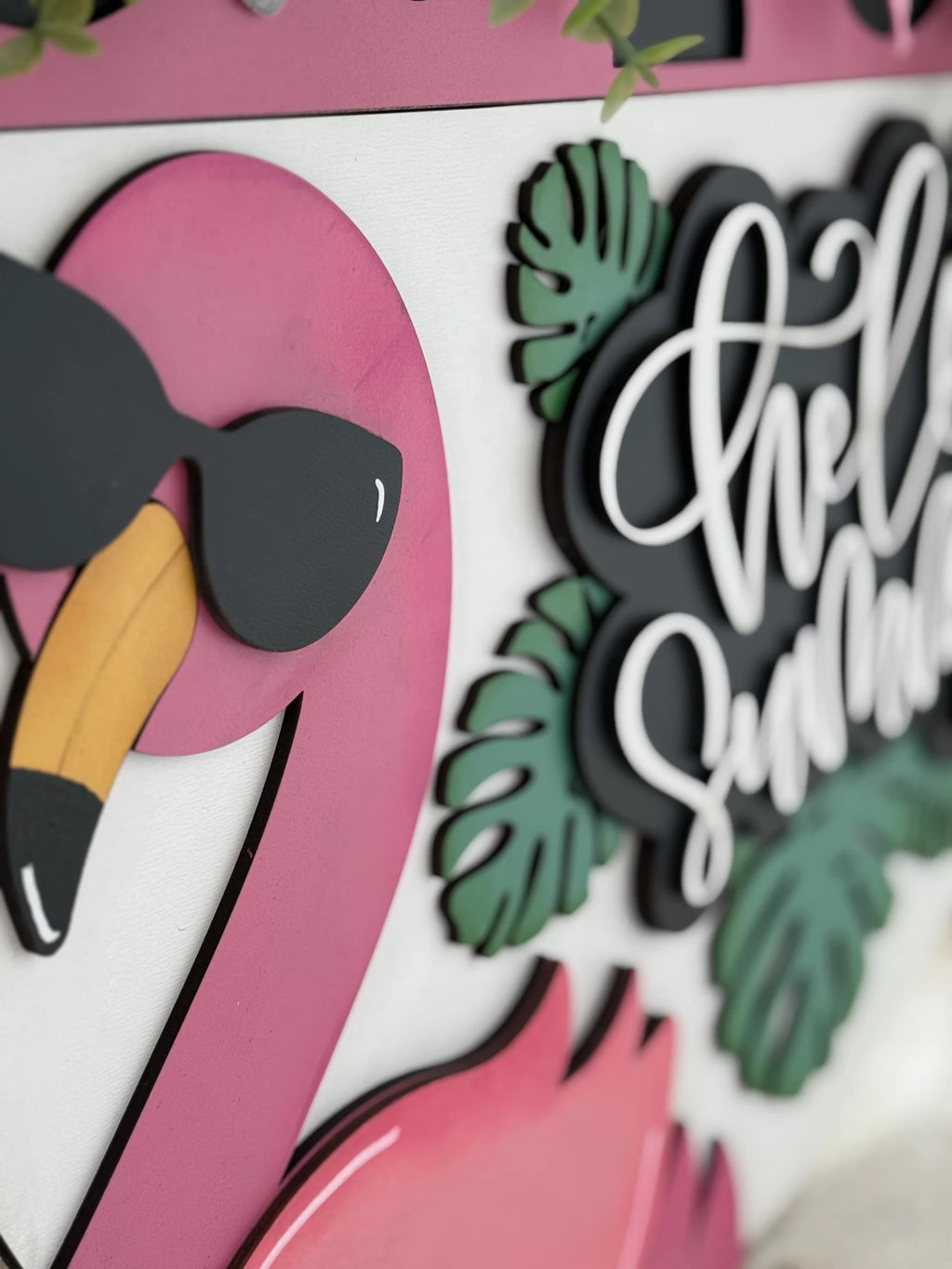 Close-up of a decorative sign featuring a pink flamingo with black sunglasses on the left. On the right, the phrase Hello Summer is written in white cursive on a black background. Green tropical leaves accent the sign, evoking a fun and summery vibe.