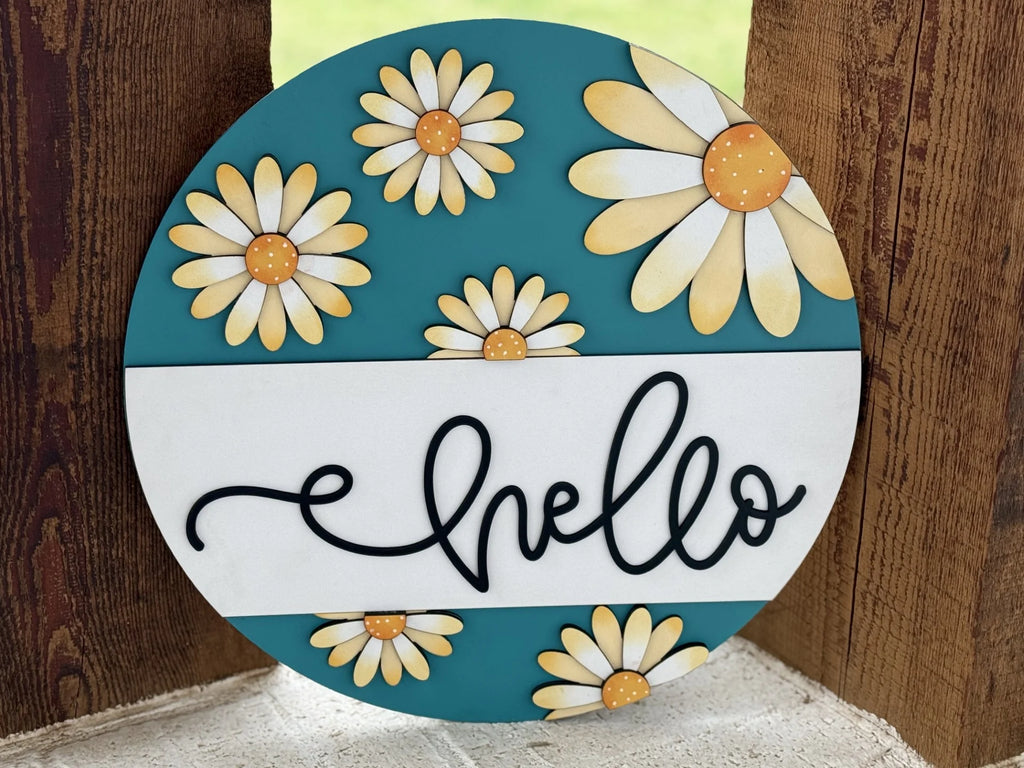 A round sign with a teal background and white daisies with yellow centers. A white horizontal stripe crosses the middle, featuring the word hello in black cursive. The sign is set against a wooden plank backdrop.