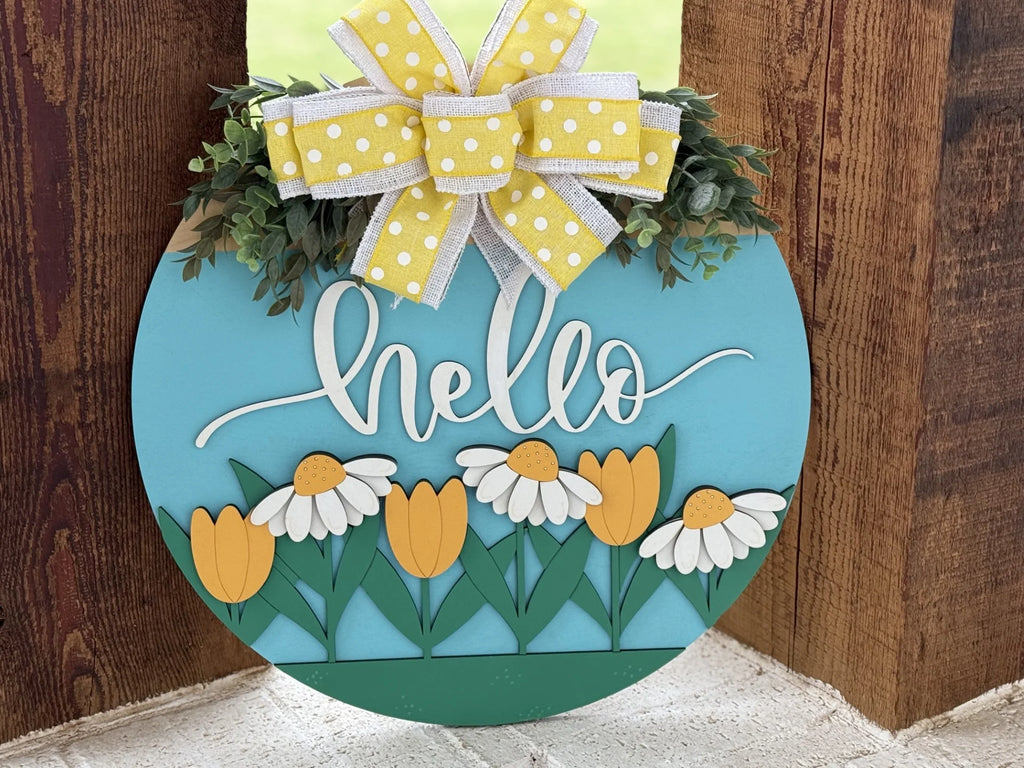 A round, light blue decorative sign with hello written in white cursive. Below, tulips and daisies are depicted. At the top, there is a yellow and white polka dot bow with greenery. The sign is set against a wooden background.