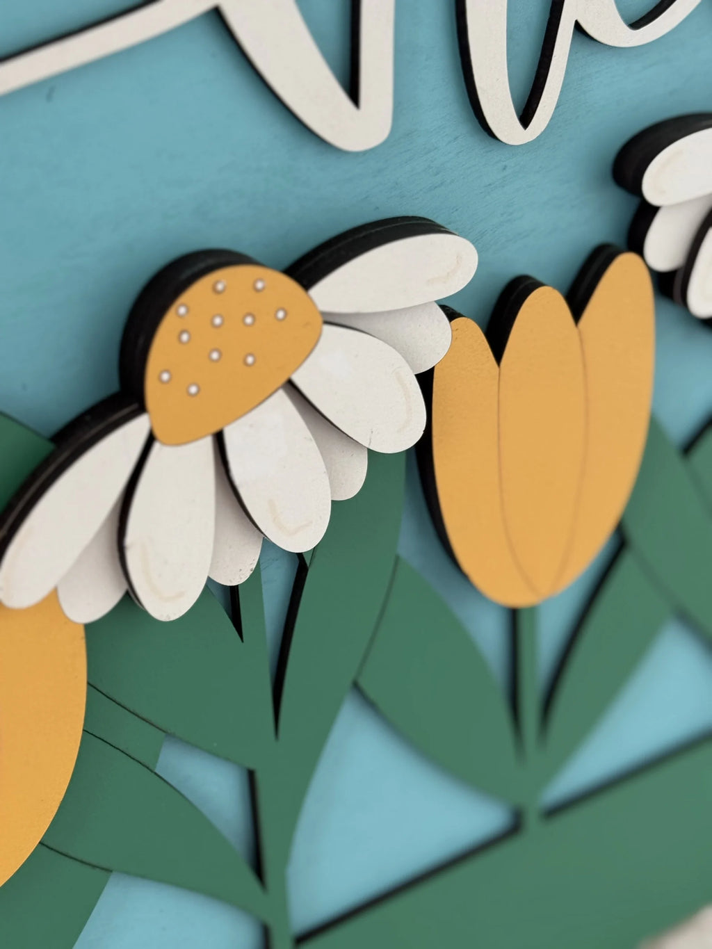 Close-up of a decorative floral piece with layered wooden cutouts. The design features a daisy with a yellow center and white petals, and yellow tulip-like flowers. Green leaves and stems are in the background, and the entire piece is set against a light blue backdrop.