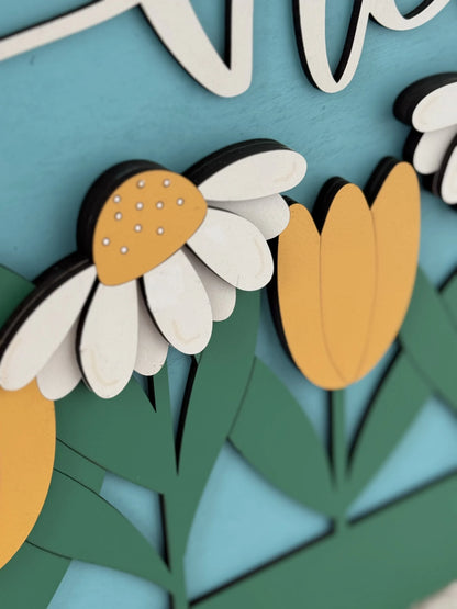 Close-up of a decorative floral piece with layered wooden cutouts. The design features a daisy with a yellow center and white petals, and yellow tulip-like flowers. Green leaves and stems are in the background, and the entire piece is set against a light blue backdrop.