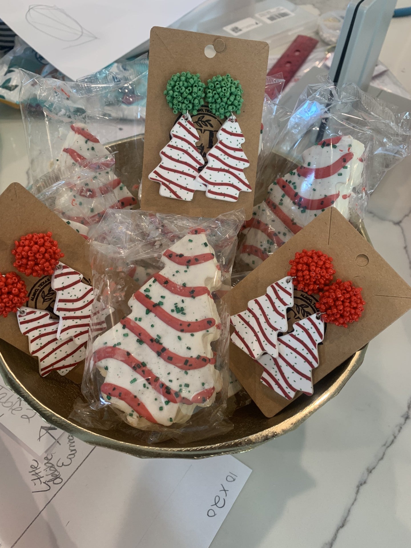 A bowl holds Little Debbie Christmas Tree Cakes Holiday Dangle Earrings on brown cards with white and red stripes and green speckles. Each card has red or green beads, and the earrings are individually wrapped in clear plastic, set against papers and a pen.