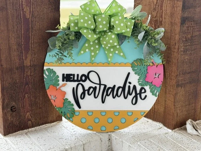 Round sign on a wooden background. The upper section features a green bow with white polka dots and green foliage. The middle has HELLO paradise in black script. The bottom is yellow with teal polka dots, accented by pink and orange tropical flowers and leaves.