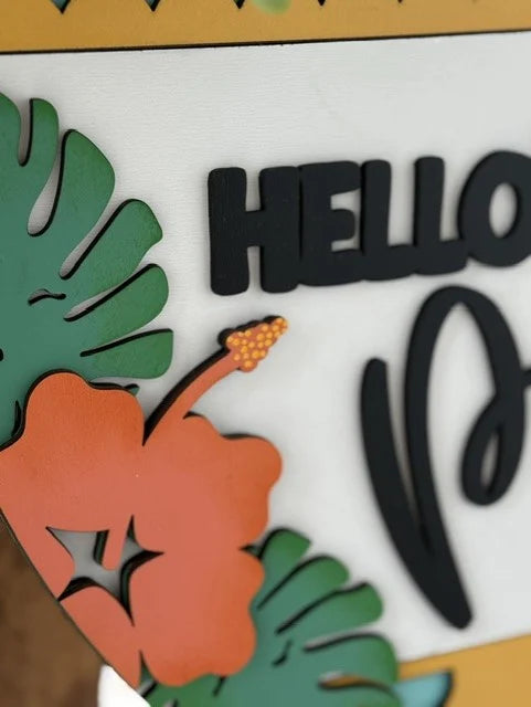 Close-up of a decorative sign featuring a colorful hibiscus flower with green leaves on the left. The word HELLO is partially visible in bold black letters on a white background. The design includes an orange and yellow palette, with a playful and tropical theme.