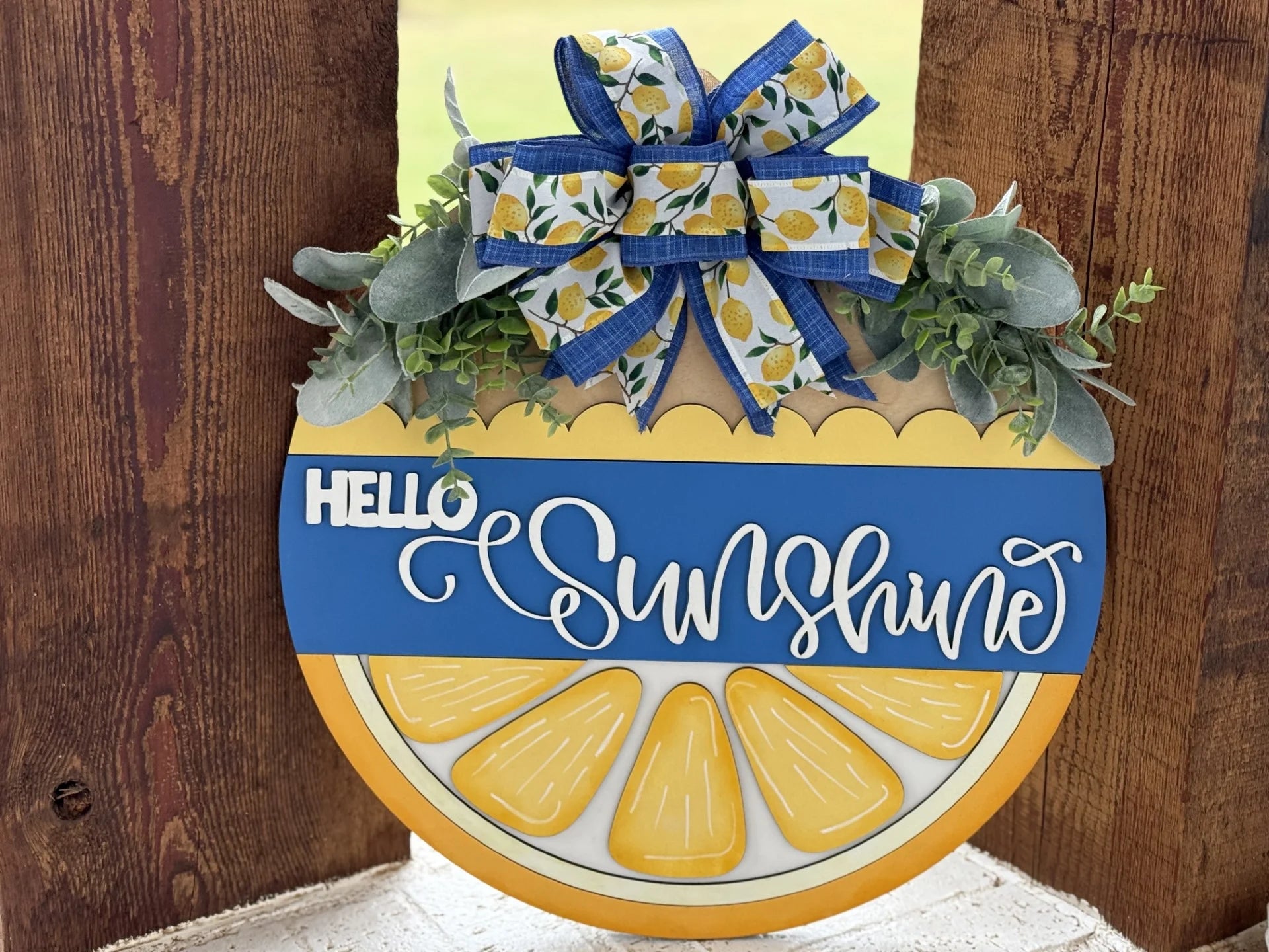 A decorative wooden sign in the shape of a lemon slice features the text Hello Sunshine in white cursive on a blue ribbon. Above, a blue ribbon adorned with lemon illustrations is tied in a bow with green foliage. The sign hangs on a wooden background.