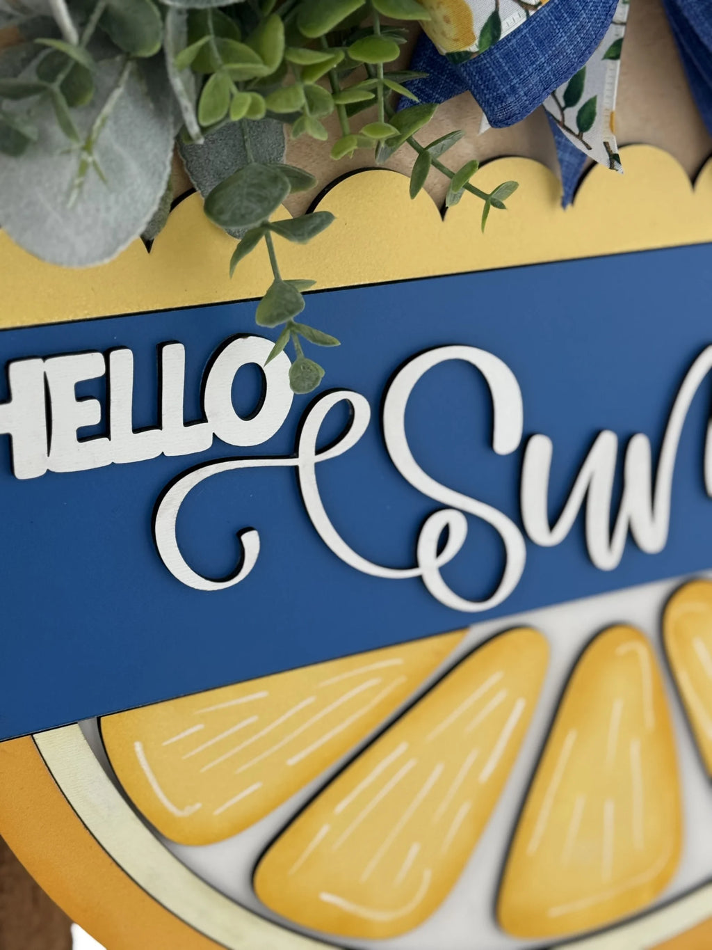 Close-up of a decorative sign featuring the words HELLO SUN in white, cursive letters on a blue background. Below are bright yellow lemon slices. Greenery and blue plaid ribbon are arranged around the top edge, adding a fresh, summery feel.