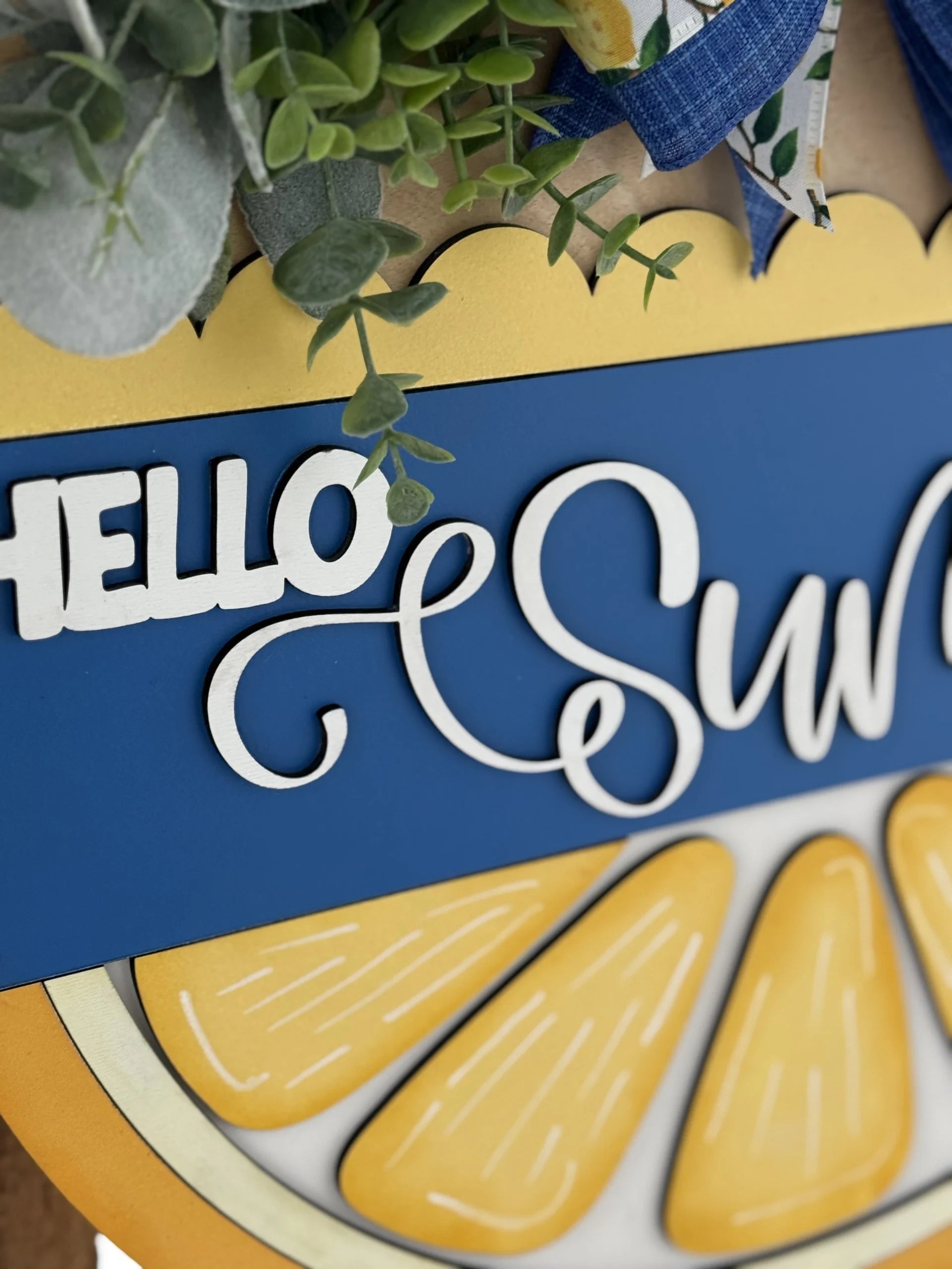 Close-up of a decorative sign featuring the words HELLO SUN in white, cursive letters on a blue background. Below are bright yellow lemon slices. Greenery and blue plaid ribbon are arranged around the top edge, adding a fresh, summery feel.